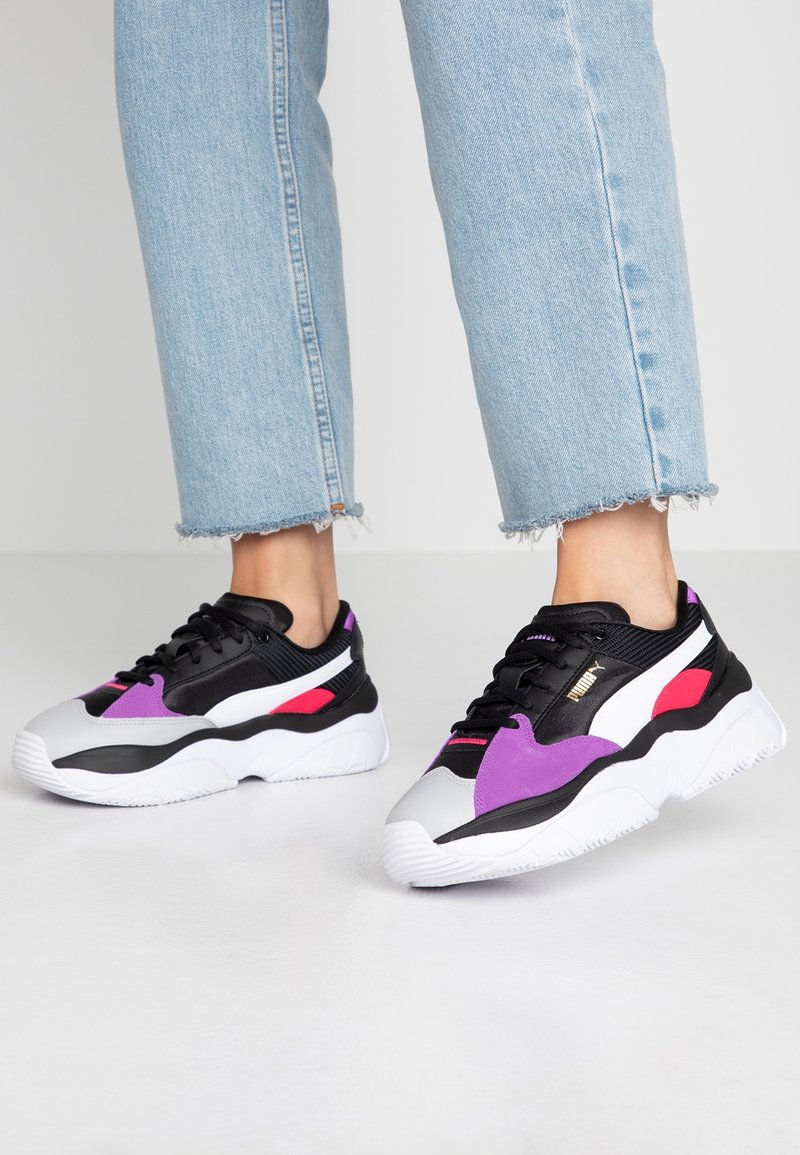 Pieds portant des baskets Puma noires, blanches, violettes et rouges, associées à un jean bleu clair court avec des ourlets effilochés, sur un sol gris.