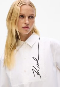 White shirt with a button-down collar, featuring a black embroidered signature detail on the chest and a smooth cotton texture.