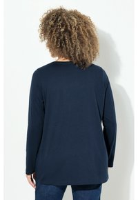 Person mit lockigem Haar, der sich abwendet, trägt ein dunkelblaues Langarmshirt und blaue Jeans vor einem schlichten weißen Hintergrund.