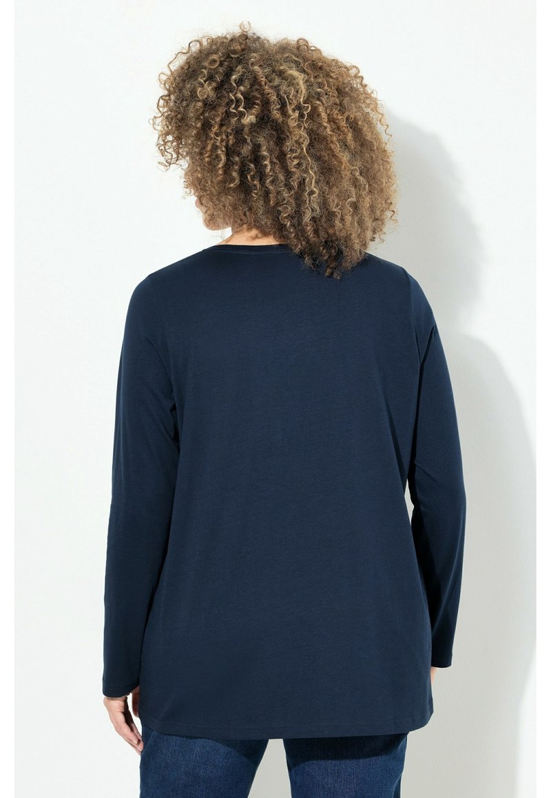 Person mit lockigem Haar, der sich abwendet, trägt ein dunkelblaues Langarmshirt und blaue Jeans vor einem schlichten weißen Hintergrund.