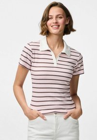 Laiši rozā krāsas polo krekls ar šaurām tumšām svītrām, balta apkakle, īsas piedurknes un pieguļošs dizains. Valkāts ar baltām biksēm.