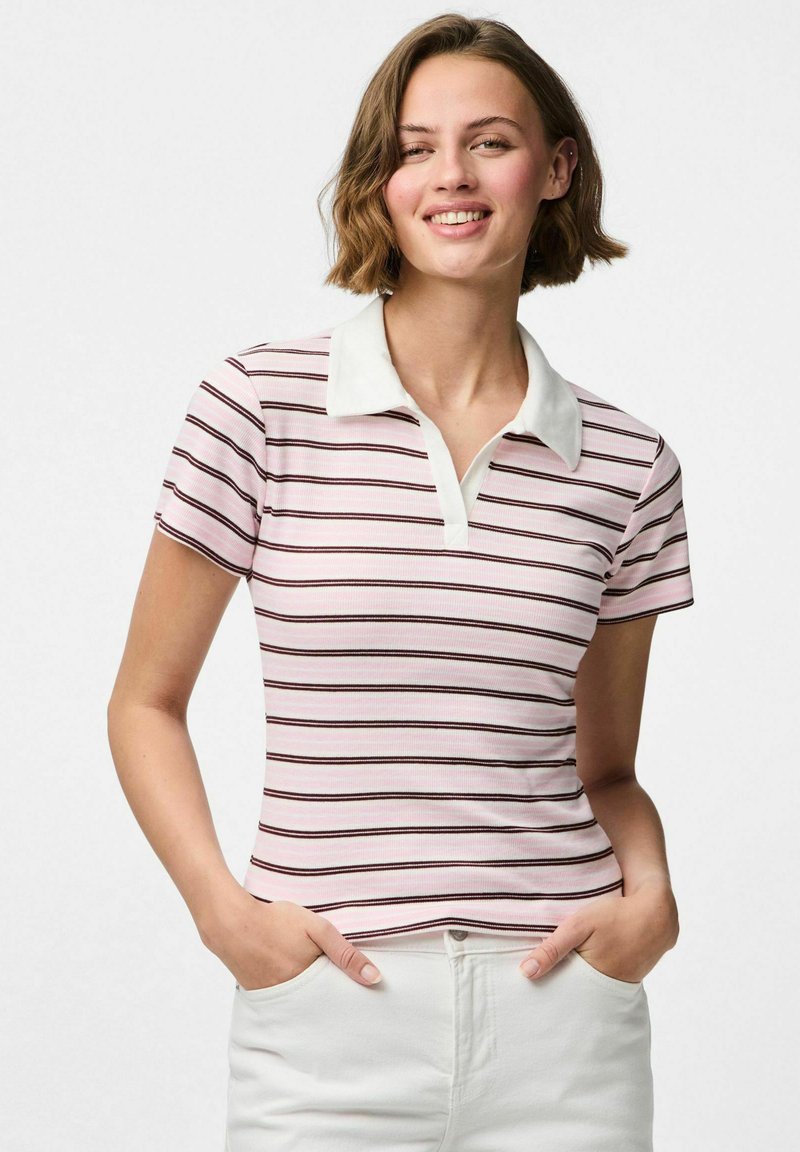 Laiši rozā krāsas polo krekls ar šaurām tumšām svītrām, balta apkakle, īsas piedurknes un pieguļošs dizains. Valkāts ar baltām biksēm.