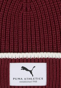 Berretto lavorato a maglia color marrone scuro con striscia orizzontale bianca. Presenta un'etichetta in tessuto con il logo Puma e la scritta "PUMA ATHLETICS fondato nel 1948."