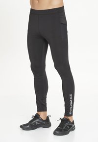 Manlig underkropp som bär svarta åtsittande leggings med sidoficka och texten "ENDURANCE" på vaden, i kombination med svarta sportskor.
