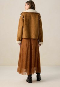 Bruine faux suède jas met schapenvachtkraag, gecombineerd met een roestkleurige kantrok en zwarte enkellaarsjes, gezien vanuit de achterkant.