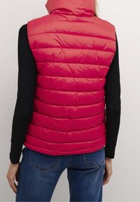 Czerwona pikowana kamizelka z wysokim kołnierzem, pikowaną fakturą i błyszczącym wykończeniem. Nosi się ją na czarnej, długorękawkowej koszuli, w połączeniu z niebieskimi dżinsami.