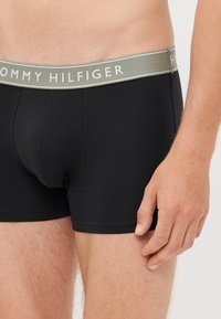 Tórax masculino e parte superior das pernas vestindo boxers pretos Tommy Hilfiger com faixa cinza na cintura, mão relaxada ao lado, tom de pele claro.