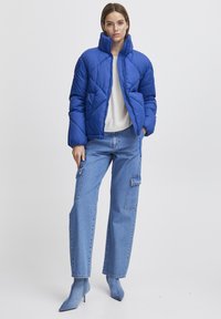 Blå puffjacka med hög krage, kviltat mönster och framzipper; kombinerad med ljusblå vida cargojeans och blå spetsiga ankelstövlar.