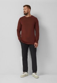 Bruin shirt met lange mouwen en ronde hals, gecombineerd met donkergrijze broek en witte sneakers, staand tegen een effen lichte achtergrond.