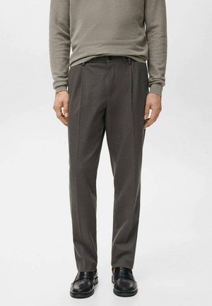 Pantalon gris sur mesure avec pli, fabriqué dans un tissu lisse. Porte un pull gris clair assorti et des mocassins noirs.