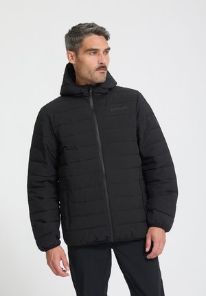 Mann mit grauen Haaren und Schnurrbart, der eine schwarze gesteppte Kapuzenjacke und schwarze Hosen trägt, schaut seitlich vor einem schlichten Hintergrund.