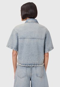 Ljusblå jeansjacka med korta ärmar, dekorerad med pärlor och silverpärlor längs fållen och en klassisk krage.