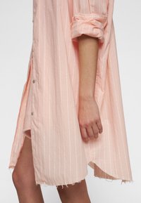 Abito camicia a righe rosa chiaro con maniche arrotolate, chiusura a bottoni, orlo irregolare e bordi sfilacciati. Tessuto morbido.