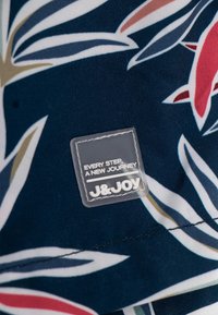 Blauwe stof met een bloemenpatroon in het wit, rood en groen. Bevat een duidelijk label met "J&JOY" en "ELKE STAP, EEN NIEUWE REIS."