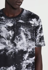 Man i svart och vitt moln-mönstrat kortärmat Puma-tröja, synlig från nacke till mitten av torso mot en enfärgad bakgrund.