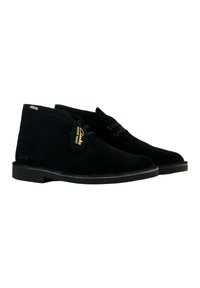 Botas de tobillo de ante negro con cordones, suela de goma negra plana, que presentan una etiqueta de marca y un detalle de costura minimalista. Diseño clásico de bota desértica.