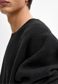 Czarny sweter wykonany na drutach o prążkowanej strukturze i szerokich rękawach, z okrągłym dekoltem i miękkim, gładkim materiałem. Zbliżenie na ramię.