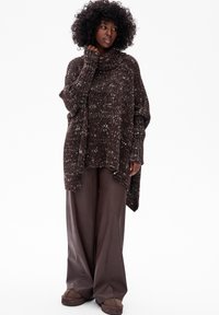 Kobieta z kręconymi włosami, ubrana w brązowy sweter typu oversize z futrzastym kołnierzem, szerokie brązowe spodnie i brązowe buty, stojąca na białym tle.