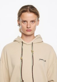 Sudadera beige con logo frontal, con cordones bicolores en marrón y azul verdoso. Tejido suave con un corte relajado y bolsillo canguro.