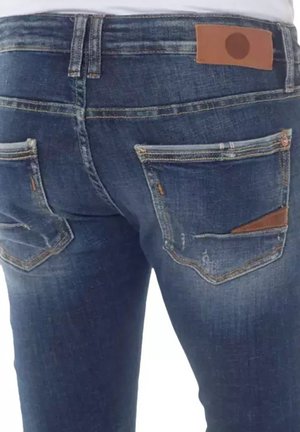Jean en denim bleu foncé avec une coupe ajustée, comportant deux poches arrière avec un accent en cuir marron sur la poche droite.