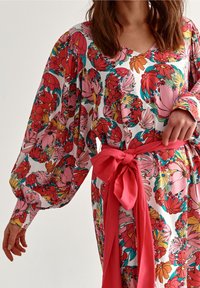 Mujer que lleva un vestido floral de mangas largas con flores rojas, rosas, amarillas y turquesas, atado en la cintura con un gran lazo de tela roja.