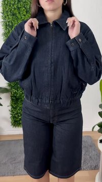Woman adjusting collar of dark denim jacket, wearing matching knee-length denim shorts, standing indoors with plants in background.