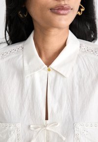 Camicia bianca con colletto, decorata con pizzo sulle spalle, un nodo in vita e un piccolo bottone dorato sul collo.
