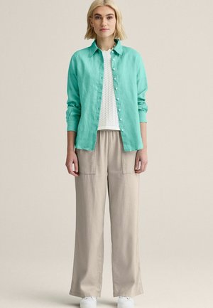 Vaaleahiuksinen nainen, joka pukeutunut turkoosiin napitettuun paitaan valkoisen neulotun topin päällä, beigen värisiin leveälahkeisiin housuihin ja valkoisiin kenkiin, seisoo yksiväristä taustaa vasten.