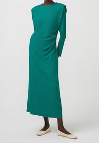 Abito lungo a maniche lunghe in teal a coste con una silhouette rilassata, dotato di spalline imbottite e drappeggi laterali. Abbinato a ballerine beige.