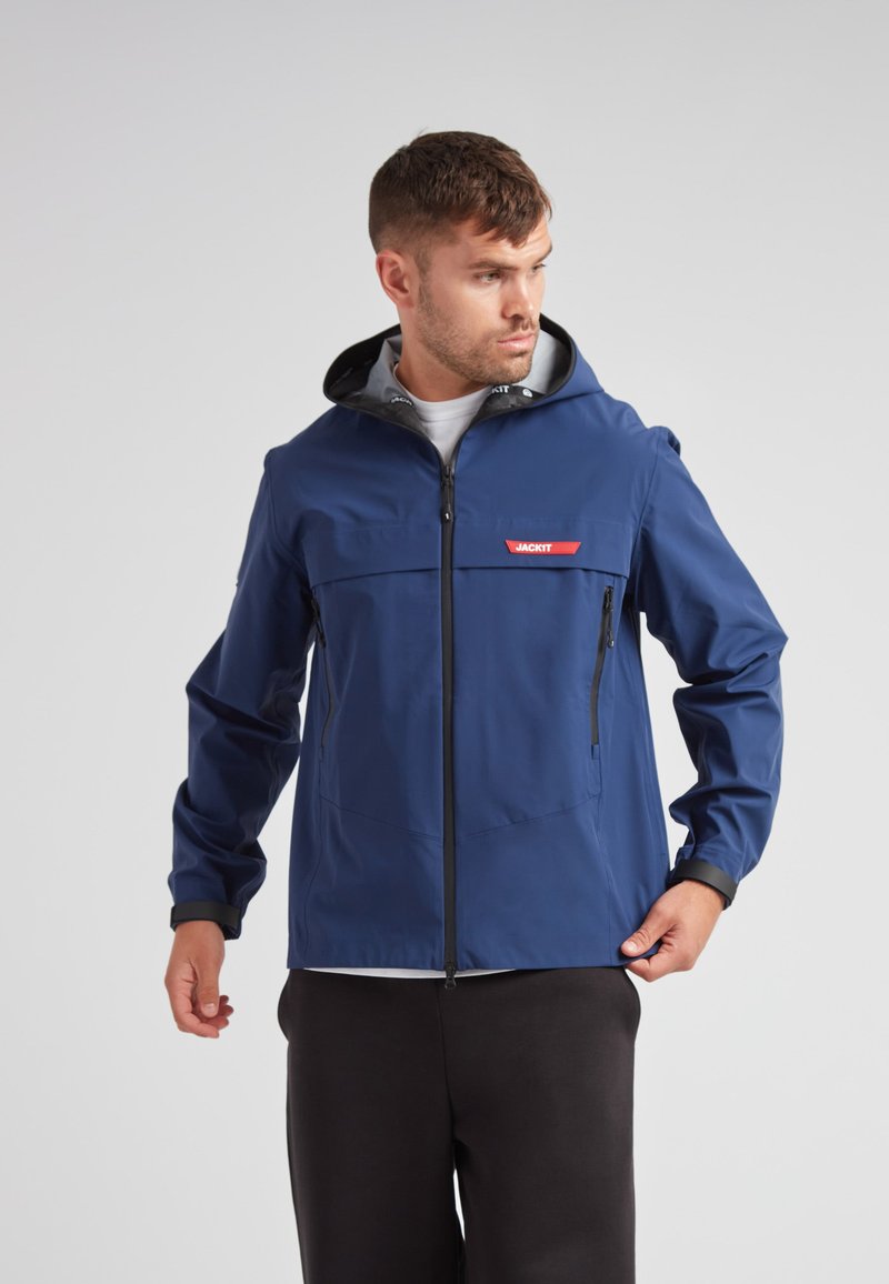 Veste imperméable bleue avec fermeture éclair à l'avant, capuche réglable, poches latérales et logo rouge contrastant. Tissu lisse et léger.