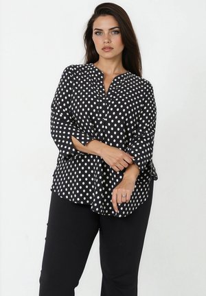 Vrouw staand tegen een witte achtergrond, gekleed in een zwart-witte blouse met polkadots en zwarte broek, met beide handen een pols vasthoudend.