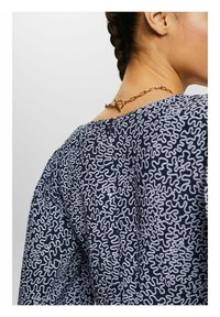 Námorně modrý top s vlnitým bílým vzorem, s kulatým výstřihem a dekorativním zlatým řetízkem na zadní straně.