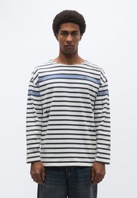 Μπλούζα μακρυμάνικη με ριγέ μοτίβο σε λευκό, μπλε και γκρι, με γραφικά τυπώματα και στρογγυλή λαιμόκοψη. Σημαντικές λεπτομέρειες περιλαμβάνουν ένα μικρό λογότυπο κορώνας.