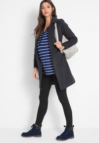 Manteau gris foncé sur un haut rayé bleu et blanc, leggings noirs et bottes bleu marine. Le modèle porte un sac gris clair, se tenant sur un fond blanc.