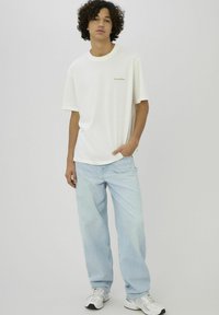 T-shirt blanc à manches courtes avec un col rond et un logo subtil, assorti à un jean bleu clair à jambes larges et des baskets blanches.