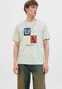 Lys grøn bomulds T-shirt med et flerfarvet grafisk tryk med abstrakte former, teksten "Original Studio" og en rund halsudskæring.