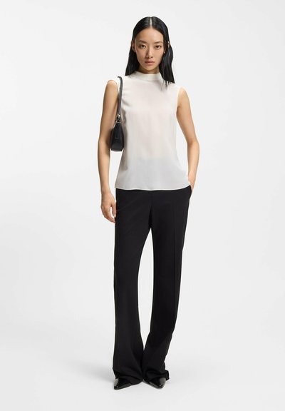 Blusa blanca transparente sin mangas con cuello alto, combinada con pantalones negros de pierna ancha. La modelo lleva un pequeño bolso negro.