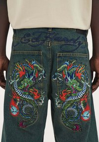 Džínové kalhoty s výšivkou zeleného draka a ohnivými akcenty. Mají tmavě modrý pas se jménem "Ed Hardy" vyšitým kurzívou.