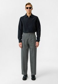 Zwarte overhemd, grijze geplooide broek en zwarte schoenen. Accessoires omvatten donkere zonnebril; zachte textuur en een getailleerde pasvorm.