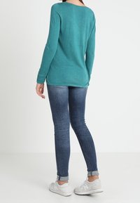 Femme portant un haut à manches longues bleu sarcelle, un jean skinny bleu foncé retroussé aux chevilles et des baskets blanches, debout devant un fond blanc uni.