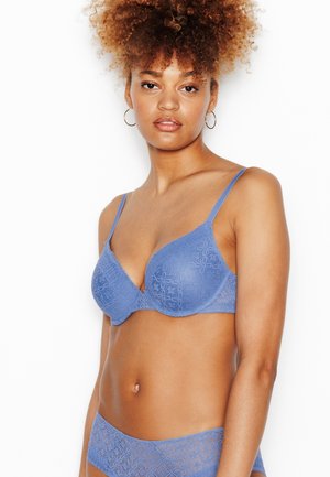 Donna con capelli ricci che indossa un reggiseno e slip di pizzo blu abbinati, in piedi su sfondo bianco, guarda avanti.