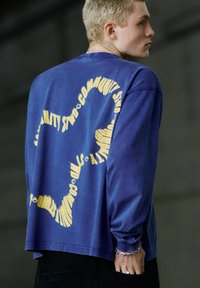 Young person wearing a blue long-sleeve shirt with a yellow wavy star-shaped design and distorted "Community Studios" text on the back.