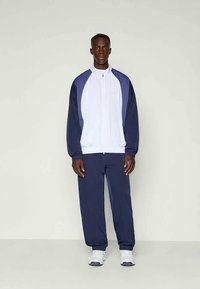 Moški, oblečen v temno modro in belo zip-up športno jakno, temno modre trenirke in bele superge, stoji ob lahkem enobarvnem ozadju.