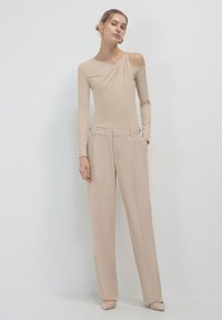 Haut beige à manches longues avec une épaule dénudée, associé à un pantalon large taille haute beige avec des accents élégants et une texture lisse.