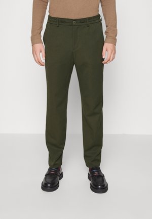 Uomo che indossa pantaloni dritti verde oliva, mocassini neri in pelle e camicia marrone a maniche lunghe, in piedi davanti a uno sfondo semplice.