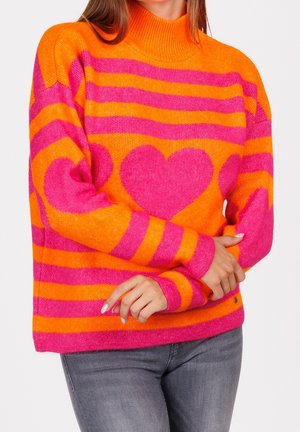 Femme portant un pull rayé orange vif et rose avec de grands motifs de cœurs roses sur la poitrine et les manches, associé à un jean gris.