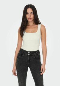 Un body de color beige claro con escote cuadrado, combinado con unos jeans negros de tiro alto que cuentan con cierre de tres botones y bolsillos, mostrando una textura suave.