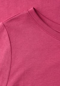 Bawełniana tkanina w kolorze fuksji, zbliżenie na zaokrąglony kołnierz o gładkiej fakturze i równych szwach wokół linii szyi i szwu ramienia.