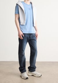 Jasnoniebieska bawełniana koszulka z małym logo, w połączeniu z ciemnoniebieskimi dżinsami. Jasnoszara bluza z kapturem zarzucona na ramiona i szare sportowe buty.