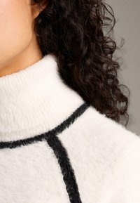 Maglione bianco a collo alto con lavorazione a maglia testurizzata, caratterizzato da accenti neri e un motivo distintivo lungo la cucitura della spalla. Materiale morbido e soffice.